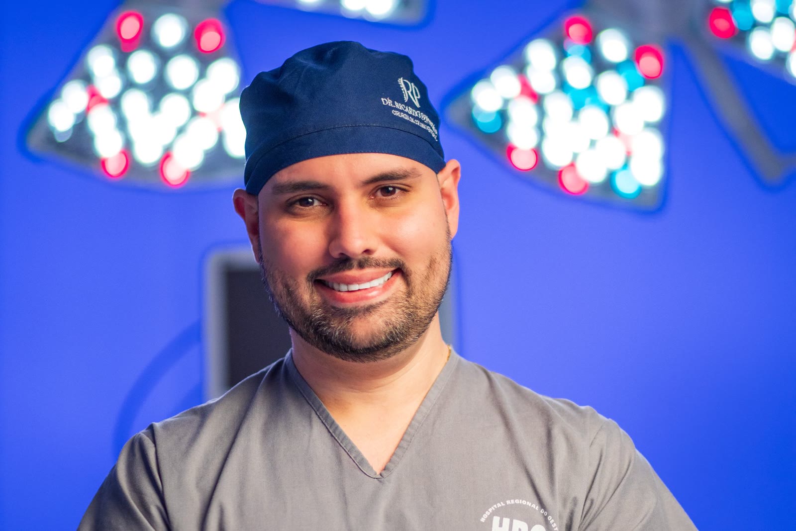 Retrato do Dr. Ricardo Penteado sorrindo, de touca cirúrgica azul, no centro cirúrgico